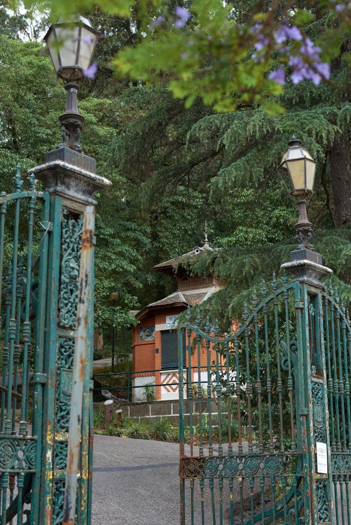 o parque de benfica é um oasis no bairro. na fotografia vemos a entrada, com os portões de ferro forjado originais e ao fundo a pequena construção de tijolo vermelho